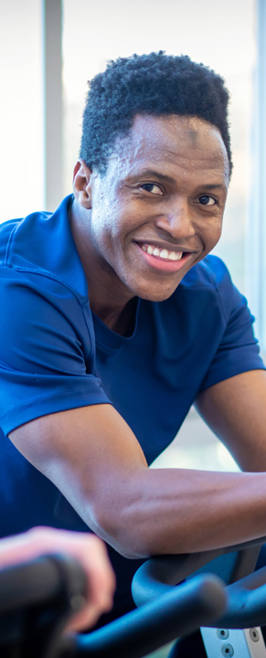 Student on a stationary bike smiling at the camera in a gym filled with natural light. Student on a stationary bike smiling at the camera in a gym filled with natural light.